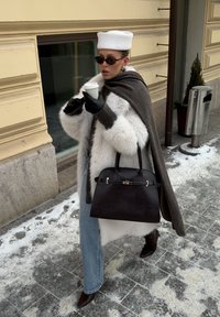 Femme en manteau de fourrure blanc, chapeau blanc et lunettes de soleil tenant une tasse de café et un sac à main noir sur un trottoir enneigé en ville.