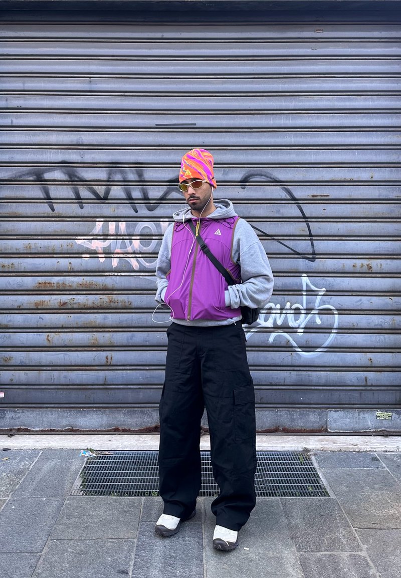 Homme portant un gilet violet par-dessus un sweat à capuche gris, un pantalon cargo noir, des baskets blanches, un bonnet à motifs orange et rose, des lunettes de soleil et des écouteurs, debout devant un rideau métallique graffité.