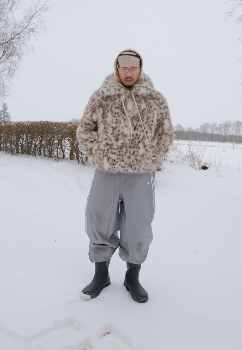 Man som bär glasögon, en prickig huva i fuskpäls, baggy grå byxor och svarta stövlar står utomhus i ett snöklätt landskap med kala träd.
