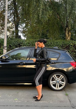 Chaqueta de cuero negra, leggings negros con rayas blancas y zapatos planos negros. De pie al lado de un coche oscuro con ruedas plateadas, con follaje verde de fondo.