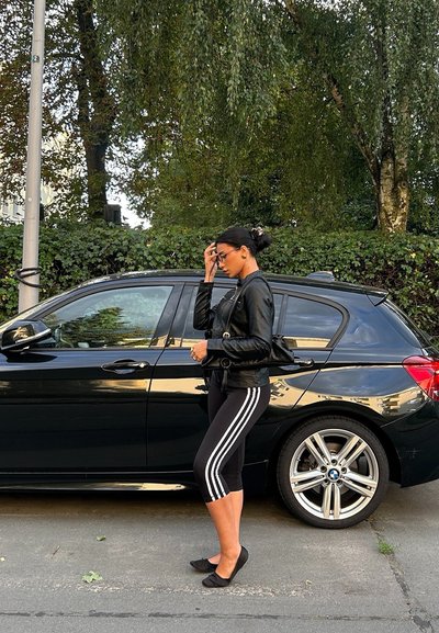 Chaqueta de cuero negra, leggings negros con rayas blancas y zapatos planos negros. De pie al lado de un coche oscuro con ruedas plateadas, con follaje verde de fondo.