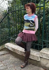 Femme aux cheveux bouclés appuyée contre une clôture, portant un gilet en tricot pittoresque, une jupe plissée mauve, des collants gris et des bottes marron montantes en plein air.