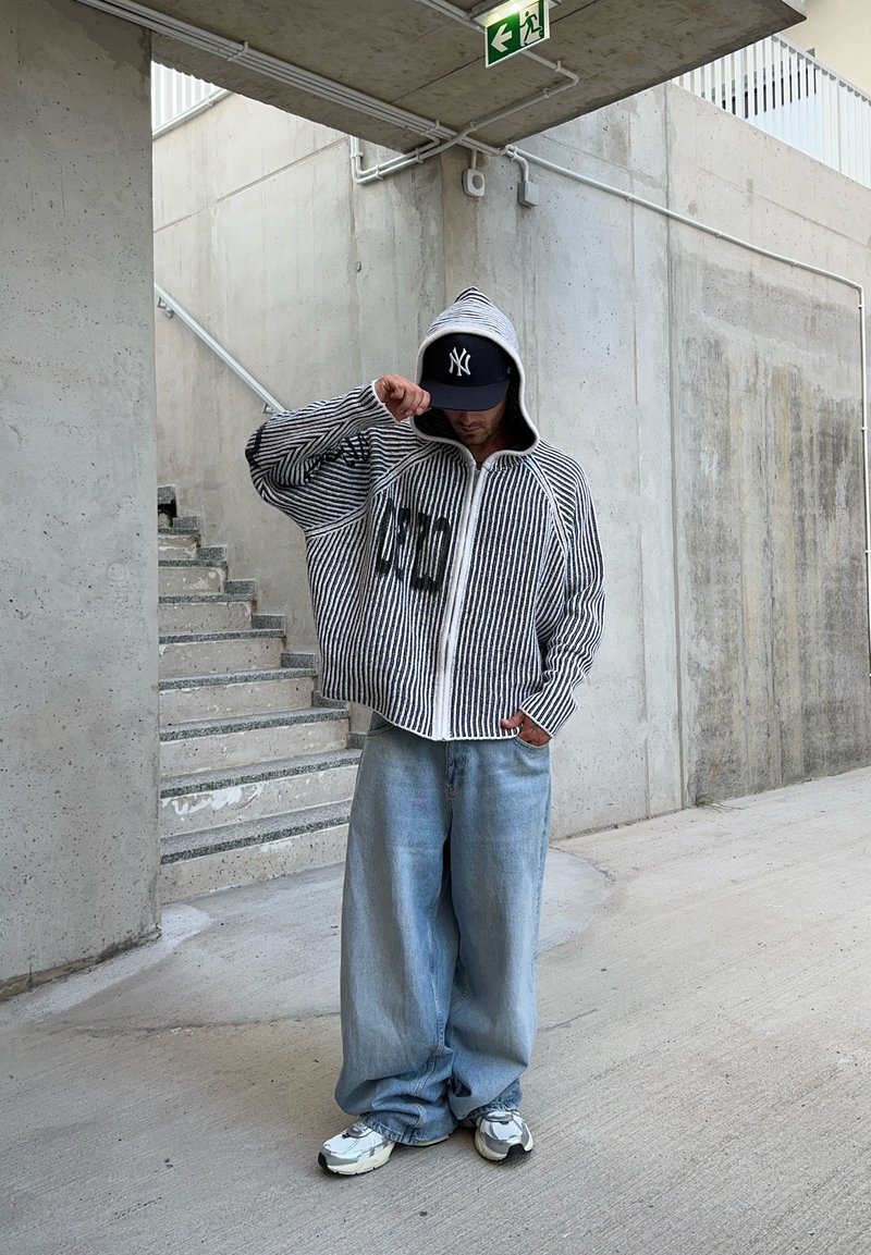 Μαύρο και λευκό ριγέ φούτερ με φερμουάρ και εφαρμοστή κουκούλα, σε συνδυασμό με ανοιχτό μπλε φαρδύ τζιν και γκρι αθλητικά παπούτσια, στέκεται σε τσιμεντένιες σκάλες.