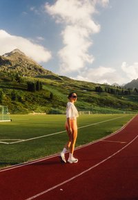 Sportiniai šortai persikų spalvos, balta įstrižinė palaidinė ir baltos sportinės bateliai. Stovi raudoname take, su žaliąja pieva ir kalnais fone.