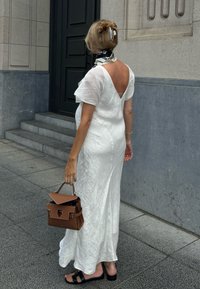 White, textured maxi dress featuring short, flutter sleeves and a V-back. Brown handbag with a top handle; black sandals with a flat sole.