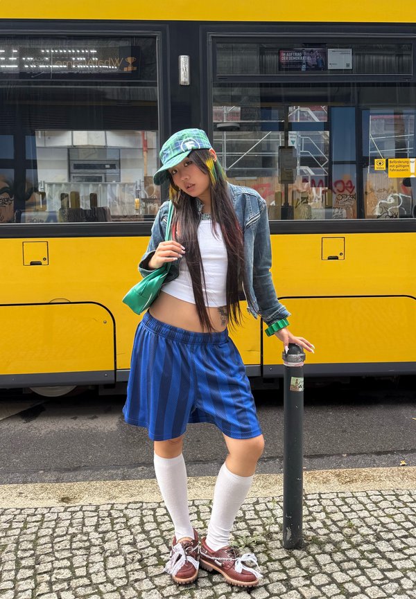 Jeune femme en short bleu à rayures, haut blanc, veste en jean, chapeau vert et chaussettes blanches montantes, debout sur des pavés près d’un tram jaune.