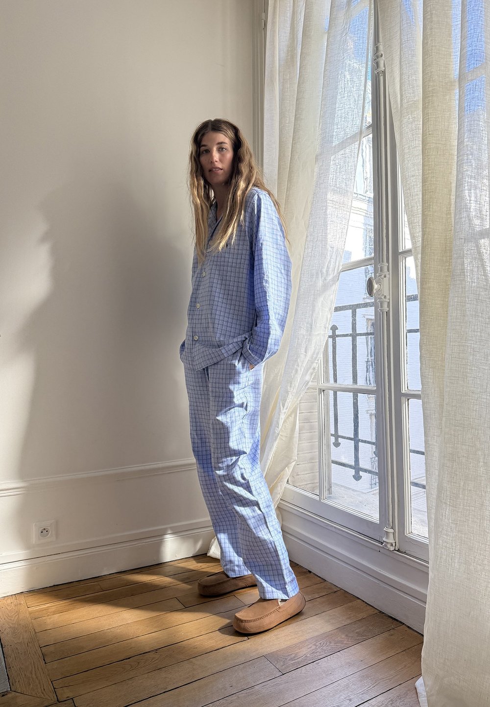 Ljusblå rutiga pyjamasset med skjorta med knappar och löst sittande byxor. Bärs med bruna tofflor. Soligt rum med vita gardiner.