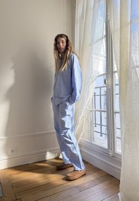 Ljusblå rutiga pyjamasset med skjorta med knappar och löst sittande byxor. Bärs med bruna tofflor. Soligt rum med vita gardiner.