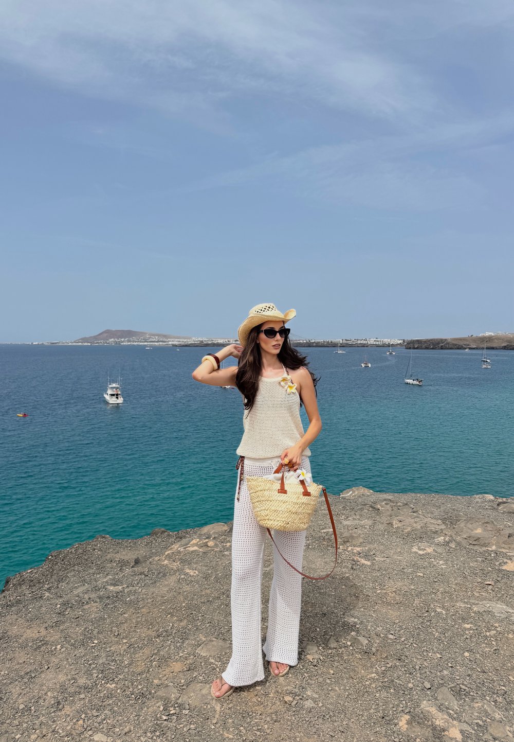 Un top din tricot subțire, de culoare crem, și pantaloni largi albi, o pălărie de paie, ochelari de soare închiși la culoare și o geantă împletită cu accente din piele maro.