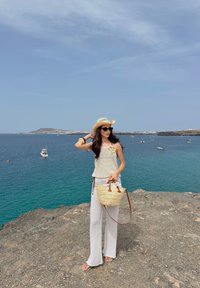Un top din tricot subțire, de culoare crem, și pantaloni largi albi, o pălărie de paie, ochelari de soare închiși la culoare și o geantă împletită cu accente din piele maro.