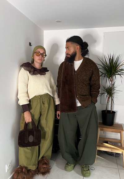 Dos personas están juntas. Una lleva un suéter de canalé color crema, pantalones verdes holgados y un bolso de mano oscuro y esponjoso. La otra lleva un cárdigan oscuro con patrón, pantalones oliva y zapatos texturizados.
