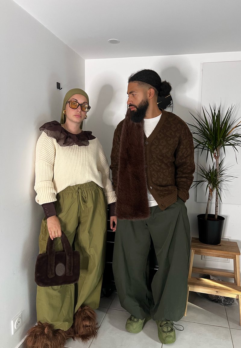 Duas pessoas estão juntas. Uma veste um suéter creme canelado, calças verdes largas e uma mala felpuda castanho-escura. A outra usa um cardigã escuro estampado, calças oliva e sapatos texturizados.