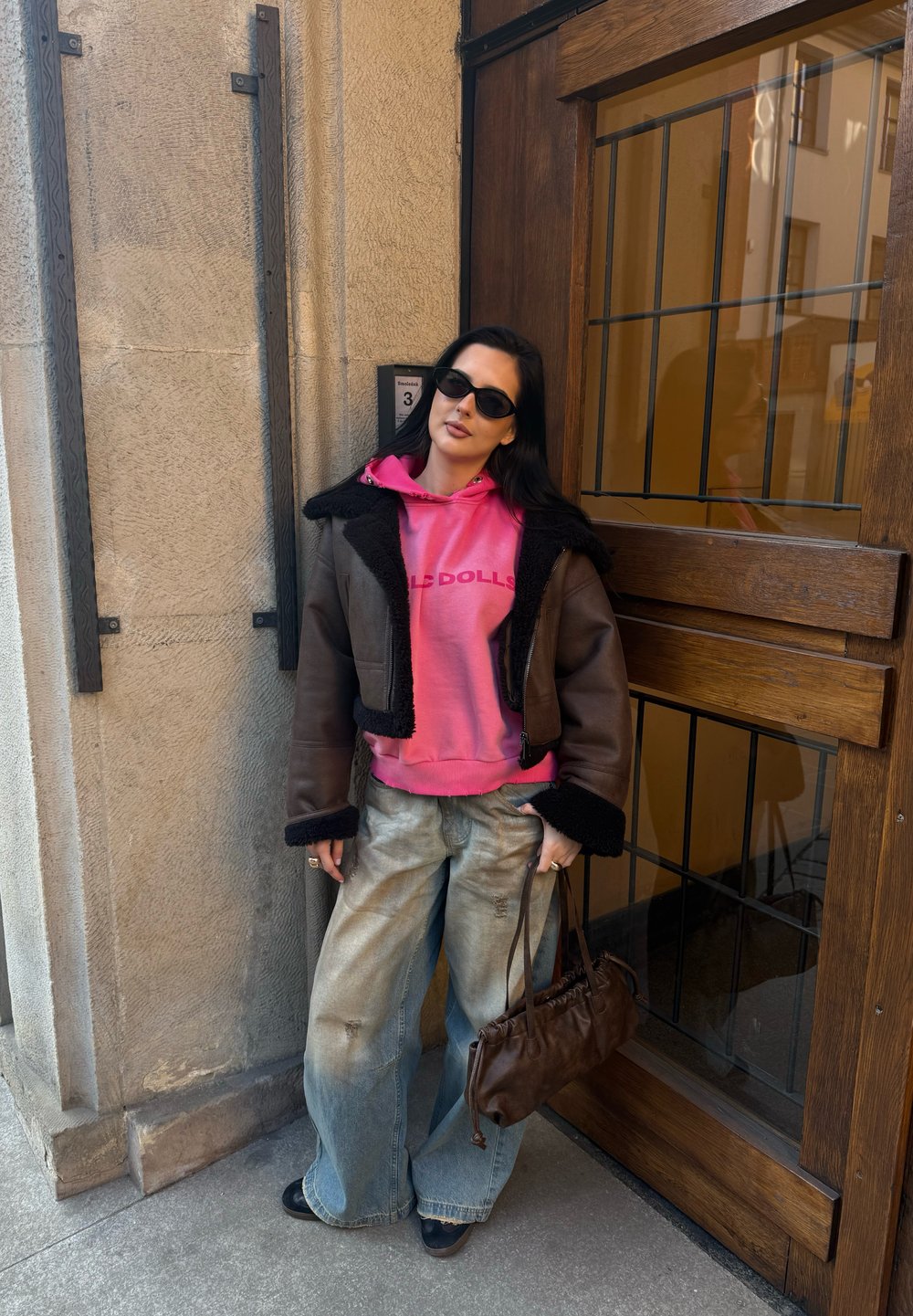Brun shearling-jakke, rosa hettegenser med tekst, vide, falmede jeans og en brun lærveske. Mørke solbriller fremhever stilen.