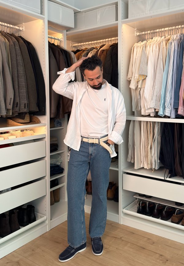 Uomo in piedi in un guardaroba ben organizzato con giacche, camicie, pantaloni e scarpe ordinatamente disposti su scaffali e appendini.