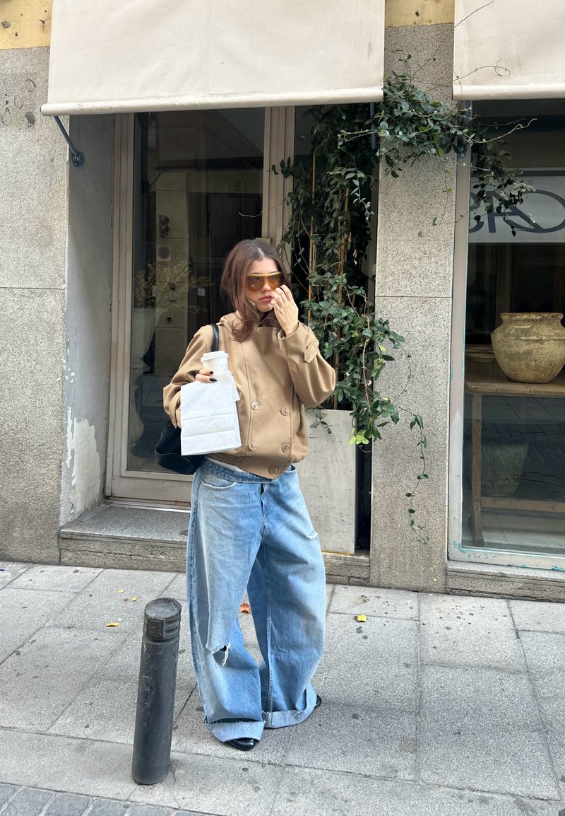 Jeune femme portant une grande veste beige et un jean large, tenant une tasse de café et un sac, debout sur un trottoir en ville près de l'entrée d'un magasin.