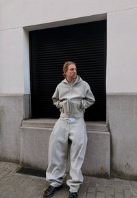 Homme en sweat à capuche gris oversize et pantalon de survêtement, les mains dans les poches, debout contre un bâtiment avec un volet roulant noir fermé.