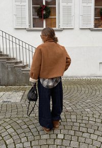 Veste marron raccourcie avec poignets en shearling, chemise à carreaux en dessous, jeans larges bleu foncé, et chaussures marron. Sac à main noir et escalier extérieur.