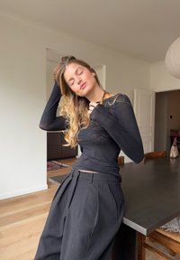 Haut noir à manches longues en matériau transparent, associé à un pantalon noir plissé taille haute. Fond d'intérieur aux tons neutres.