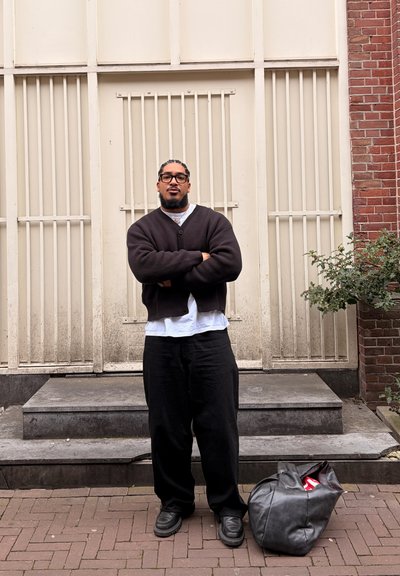 Hombre con gafas y cabello trenzado está de pie con los brazos cruzados delante de una puerta blanca con barrotes, bolsa negra en el suelo cerca.