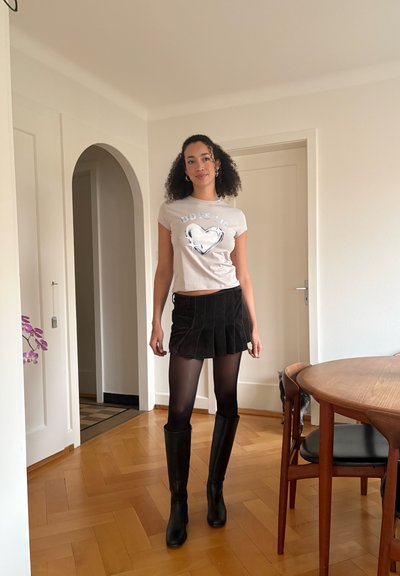 Mujer con cabello rizado de pie en interiores, lleva una camiseta blanca con diseño gráfico, minifalda negra plisada, medias negras y botas negras hasta la rodilla.