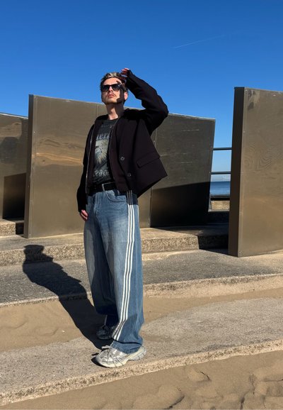 Chaqueta negra tipo cropped, camiseta gráfica, vaqueros anchos de mezclilla azul con franjas laterales y zapatillas plateadas. Persona posa al aire libre contra paneles de metal lisos.