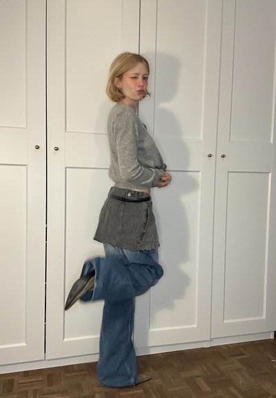 Suéter gris, falda de mezclilla negra superpuesta sobre jeans sueltos de color azul claro, con bordes deshilachados; pose de pie frente a puertas de un armario blanco.