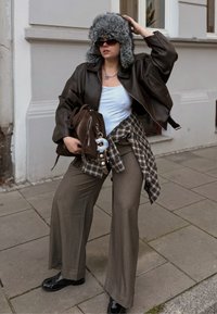 Giacca corta in pelle marrone, canottiera bianca, camicia a quadri grigia annodata in vita, pantaloni larghi marroni, scarpe nere lucide, cappello di pelliccia sintetica grigio, occhiali da sole.