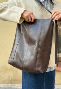 Mains avec des bagues tenant un sac à bandoulière en cuir brun foncé ouvert, porté sur un pull beige et une chemise rayée bleue, sur fond de jupe en denim.