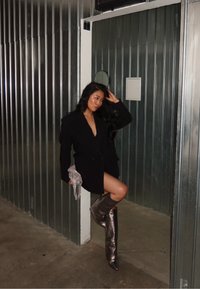 Skinnen sølvfargede støvler med spisse tær som går opp til knærne, brukt sammen med en svart overdimensjonert blazer. Bakgrunnen har metallpaneler og et speil.