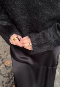 Pull noir et duveteux à manches longues avec des poignets côtelés ; porté sur une jupe en satin noir brillante. Deux bagues avec des accents dorés aux mains.