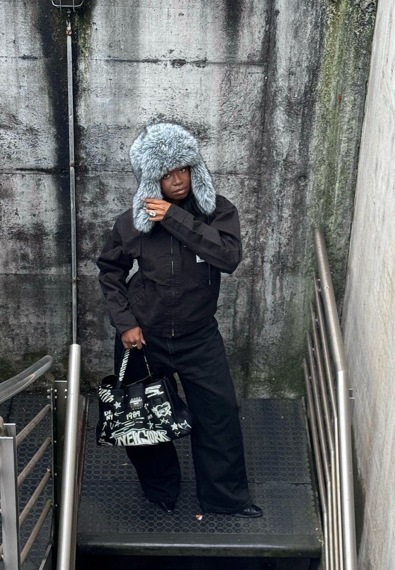 Personne en veste et pantalon noirs portant un chapeau en fourrure grise, tenant un sac noir style graffiti, debout sur des escaliers métalliques contre un mur de béton texturé.