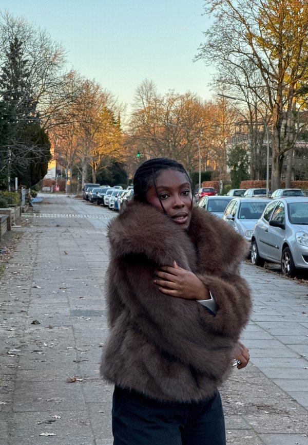 Cappotto in pelliccia sintetica marrone con collo ampio, design corto e texture morbida, indossato sopra una maglia bianca e pantaloni neri, in un ambiente urbano all'aperto.