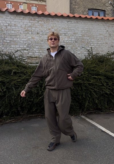 Joven con sudadera marrón holgada y pantalones, zapatillas oscuras y gafas de sol, de pie en una zona pavimentada con muro de ladrillos y arbustos detrás.