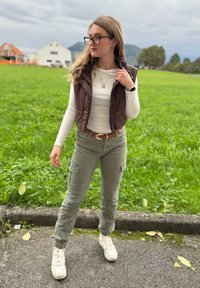 Brun vadderad väst över en vit långärmad skjorta, olivgröna cargobyxor med fickor och vita sneakers. Texturerad gräsmatta i bakgrunden.