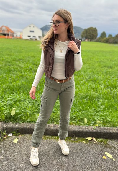 Chaleco acolchado marrón sobre una camisa de manga larga blanca, pantalones cargo oliva con bolsillos y zapatillas blancas. Fondo de hierba texturizada.