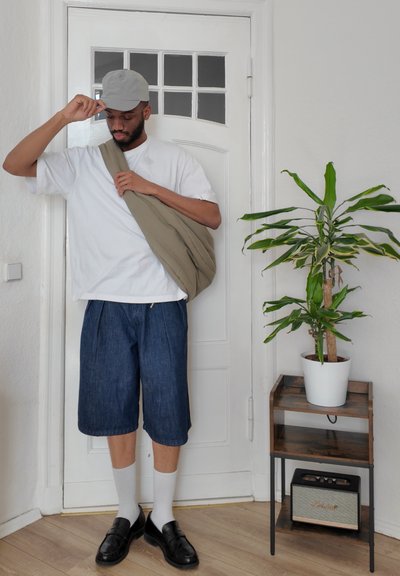 Conjunto casual que incluye una camisa blanca, pantalones cortos de mezclilla, una gorra gris, un bolso de hombro beige, mocasines negros, calcetines blancos y una planta en maceta cercana.