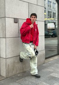 Κόκκινη κουκούλα κάτω από ένα ταιριαστό τζάκετ, φθαρμένα ανοιχτό μπλε τζιν και μαύρα παπούτσια. Άτομο κρατά ακουστικά, ακουμπισμένο σε έναν τοίχο.