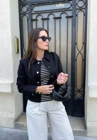 Černá džínová bunda se stříbrnými knoflíky přes černobílý pruhovaný top, spárovaná s bílými širokými kalhotami. Drží černou pletenou kabelku.