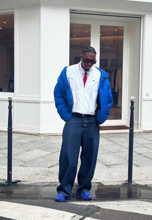 Hombre con chaqueta acolchada azul, camisa blanca con cremallera y corbata roja, vaqueros anchos oscuros y zapatillas azules de pie en una acera de la ciudad.