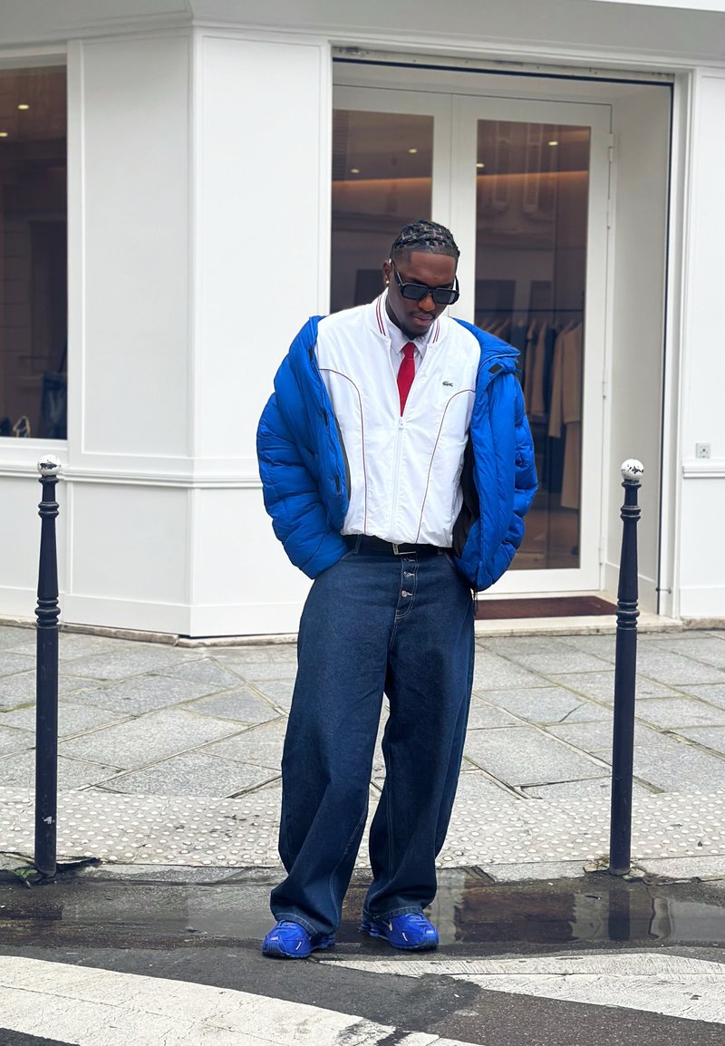 Mann trägt blaue Steppjacke, weißes Reißverschlusshemd mit roter Krawatte, weite dunkle Jeans und blaue Turnschuhe, steht auf einem Bürgersteig in der Stadt.