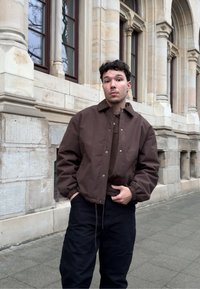 Νέος άνδρας που φοράει καφέ μπουφάν και μαύρο παντελόνι στέκεται σε πεζοδρόμιο με πλακόστρωτο δίπλα σε ιστορικό κτίριο με αψιδωτά παράθυρα.