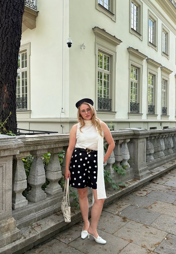 Mladá žena nosí čierny baret, biele tielko bez rukávov, čierne krátke nohavice s bodkami, biele topánky a drží béžovú tašku, stojí pri kamennom zábradlí vonku.