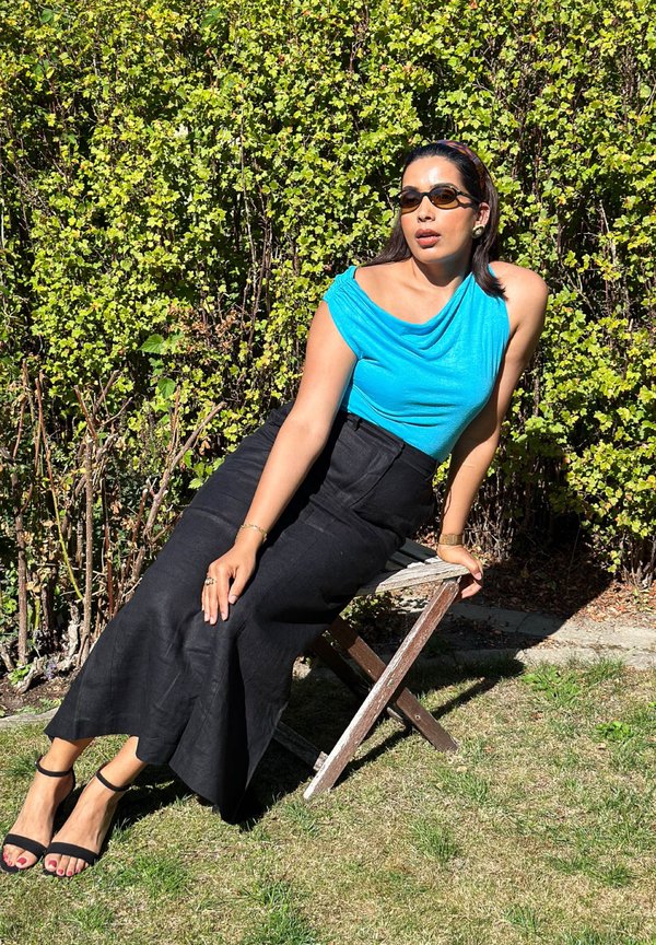 Femme portant un haut bleu, une jupe noire et des lunettes de soleil assise sur une chaise en bois à l'extérieur avec des buissons feuillus verts en arrière-plan.
