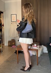 Γυναίκα με μακριά κυματιστά μαλλιά, ντυμένη με μαύρο δερμάτινο μπουφάν, λευκή Πλισέ φούστα και καφέ γόβες, στέκεται σε σαλόνι κρατώντας μια μπλε τσάντα με καρφιά.