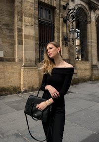 Top nero a coste scollato sulle spalle, pantaloni a vita alta coordinati e una borsa nera strutturata. Accenti di gioielli in oro. Sfondo di muro in pietra.