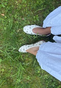 Białe skórzane buty wsuwane z okrągłymi, srebrnymi ćwiekami wzdłuż brzegów, leżące na zielonej trawie, z przewiewnym materiałem obok.