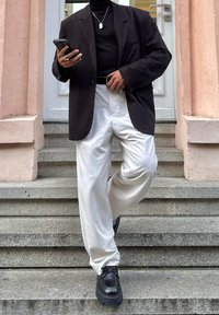 Blazer nero sopra un dolcevita nero, pantaloni bianchi a gamba larga e scarpe nere chunky. La figura tiene uno smartphone mentre è in piedi su gradini di pietra.