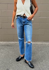 Κρέμα ριγωτό πλεκτό γιλέκο με V-λαιμό, μπλε ψηλόμεσα τζιν με σκισίματα στα γόνατα και μαύρες λόφερς με φουντάκια. Περιλαμβάνεται μαύρη ζώνη.