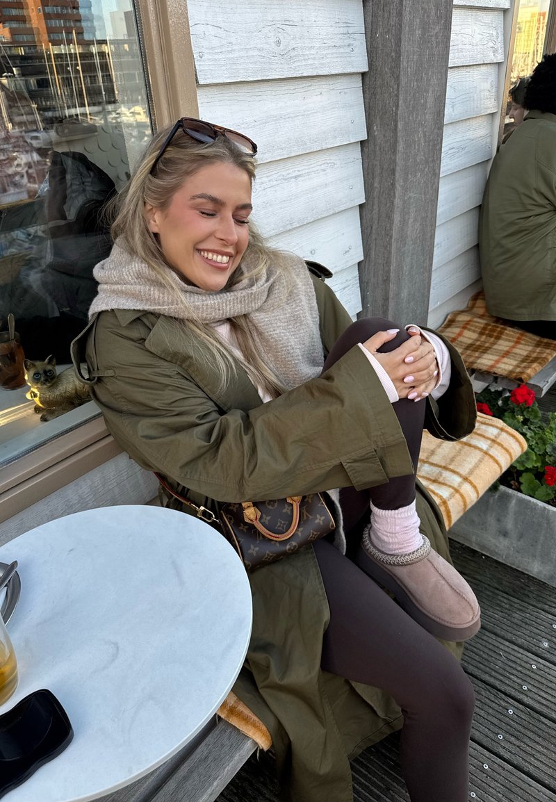 Kvinne i grønn kåpe og skjerf sitter smilende på en utendørs benk med beina i kors, solbriller på hodet, nær et bord med drikker og en kattefigur.