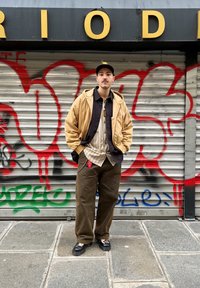 Καφέ κουκούλα πάνω από σκούρο πουκάμισο με γιακά, ριγέ πουκαμισάκι, φαρδύ καφέ παντελόνι, μαύρα παπούτσια με διακοσμητικές λεπτομέρειες, μπροστά σε γκραφίτι.